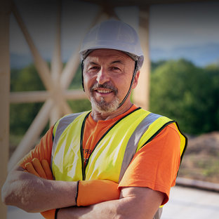 Holzbau & Holzleimbau Lächelnder Bauarbeiter mit weißem Schutzhelm und Warnweste vor einem Holzrahmenbau
