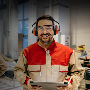 Holz- & Möbelindustrie Lächelnder Mitarbeiter mit Gehörschutz und Schutzbrille hält ein Tablet in einer Werkhalle